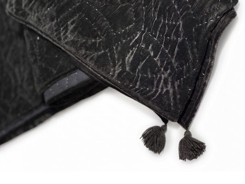 Puede incluir: Una manta plegada de terciopelo negro con un patr&oacute;n acolchado y dos borlas. La manta tiene un aspecto desgastado con costuras blancas y un borde gris oscuro. Las borlas son de un color gris oscuro a juego.