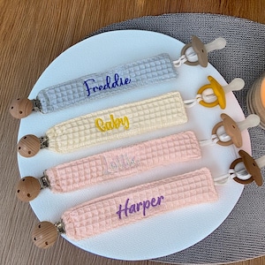 May include: Four baby pacifier clips with names: "Freddie", "Baby", "Lollie", and "Harper". The clips are in light blue, yellow, and pink waffle fabric. Each clip has a wooden clip and a pacifier attached. The pacifiers are in various colors.