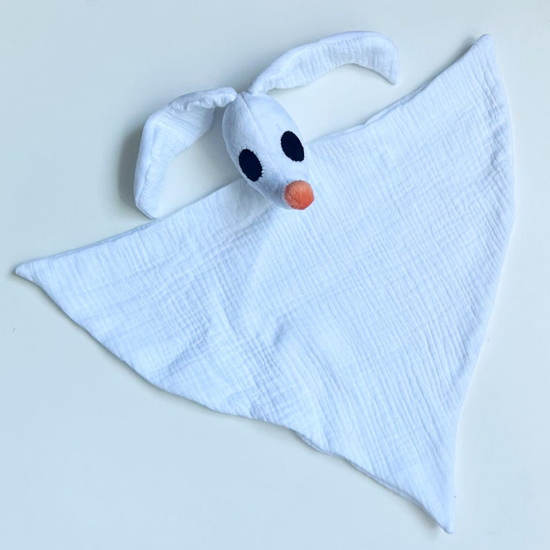 Op de afbeelding: Een witte pluche spook-knuffeldoekje met een lichaam van zachte, getextureerde mousseline stof. Het hoofd heeft twee lange, slappe oren, twee grote zwarte ovale ogen en een kleine, ronde oranje neus, lijkend op een spookachtig hondenfiguur.