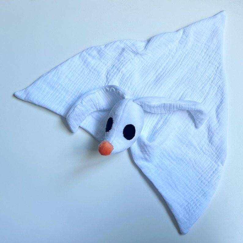 Op de afbeelding: Een wit mousseline knuffeldoekje met een pluche spookachtig karakterhoofd met lange slappe oren, grote zwarte ovale ogen en een kleine oranje neus. De stof heeft een gekreukelde textuur en is gevormd tot een zacht, driehoekig doekje.
