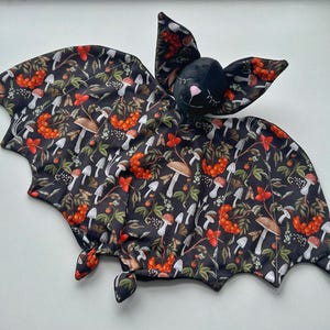 May include: A plush bat-shaped toy with a black body and wings. The wings feature a dark floral pattern with mushrooms, berries, and leaves in orange, white, and brown. The bat's head is black with closed eyes and pink cheeks.