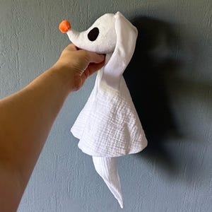 Op de afbeelding: Een wit pluche spookhondspeeltje met lange slappe oren, een kleine ronde oranje neus en grote zwarte ovale ogen. Het lichaam is gemaakt van zachte, getextureerde mousseline stof die naar beneden valt en een vloeiend silhouet cre&euml;ert.