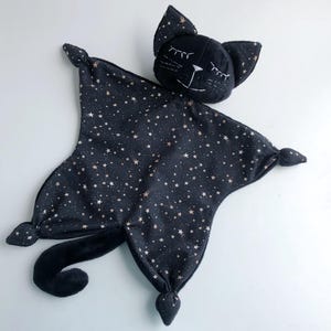 May include: A black and white cat-shaped baby security blanket. The blanket is dark blue with a pattern of small, gold-colored stars. The cat's head has closed eyes and embroidered whiskers. The tail is a soft, black plush.