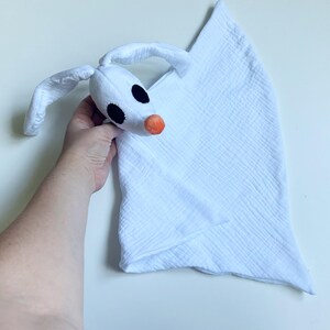 Op de afbeelding: Een wit pluche knuffeldoekje met een spookachtig hoofd, lange slappe oren, zwarte ronde ogen en een kleine oranje neus. Het bijbehorende doekje is gemaakt van zachte, gekreukelde mousseline stof in een driehoekige vorm.