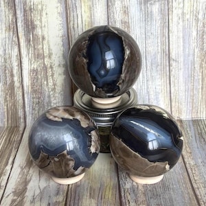 May include: Three polished agate spheres with a mix of blue, brown, and cream colors. The spheres have a glossy finish and sit on small, light-colored bases. One sphere is cut open, revealing a blue interior.