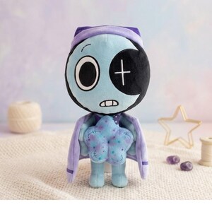 Pode incluir: Boneca de pel&uacute;cia com cabe&ccedil;a e corpo azul claro, usando capa e chap&eacute;u roxos. A boneca tem um olho preto com uma cruz branca e segura uma almofada azul em forma de estrela. Um item artesanal e fantasioso.
