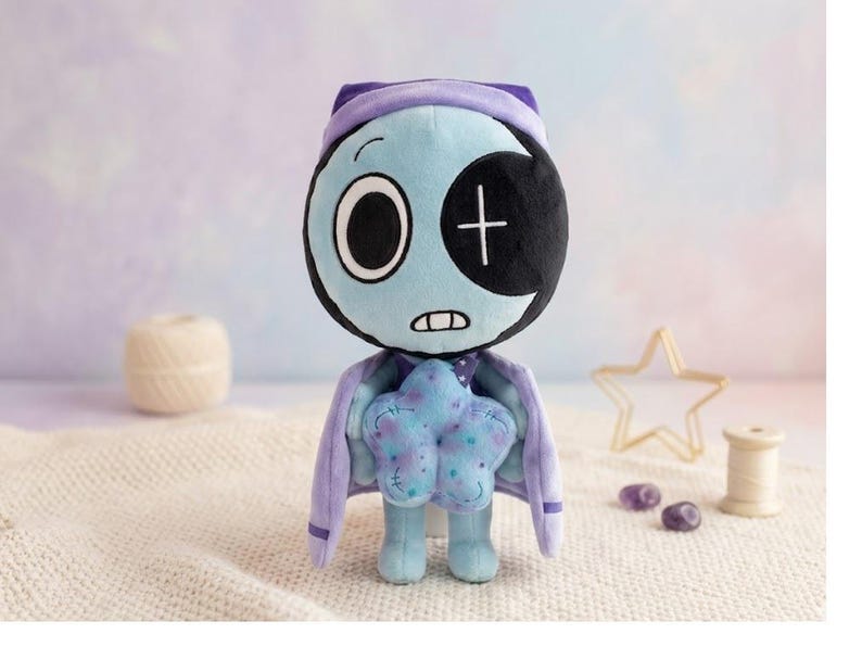 Pode incluir: Boneca de pel&uacute;cia com cabe&ccedil;a e corpo azul claro, usando capa e chap&eacute;u roxos. A boneca tem um olho preto com uma cruz branca e segura uma almofada azul em forma de estrela. Um item artesanal e fantasioso.