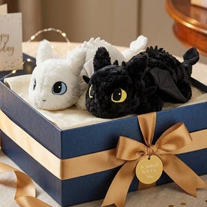 Könnte beinhalten: Eine marineblaue Geschenkbox mit einem weißen und schwarzen Drachen-Plüschtier. Die Box ist mit einem goldenen Band und einem Anhänger mit der Aufschrift "A Special Gift For You" verziert. Eine Karte mit "Happy Birthday" ist ebenfalls enthalten.