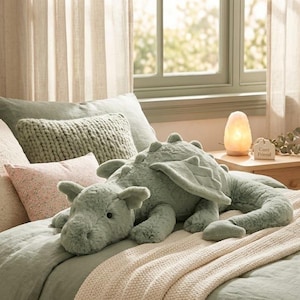 Puede incluir: Un peluche de dragón verde claro con alas y una larga cola descansa sobre una cama con ropa de cama verde salvia. La cama está adornada con varias almohadas en tonos rosas y verdes, creando un ambiente acogedor y acogedor.