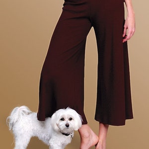 May include: A white dog with a black collar stands in front of a person wearing brown wide-leg pants.