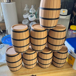 Puede incluir: Una colección de pequeños recipientes de madera con forma de barril. Cada barril es de color marrón claro con bandas negras alrededor del centro. Los barriles están apilados.