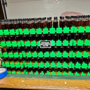 Może przedstawiać: Prezentacja małych szklanych butelek wypełnionych bursztynowym płynem, ułożonych w wielopoziomowym, zielonym plastikowym stojaku. Butelki mają czarne zakrętki. Znak głosi "INDIANA BOURBON CLUB".