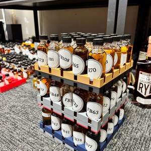 May include: A display of small liquor bottles, arranged in a cube formation. Each bottle has a white label with black text. The bottles are filled with amber-colored liquid. The display is on a gray carpet.