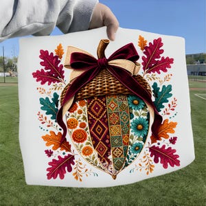 May include: A white fabric square featuring a central acorn illustration with a woven cap, a large burgundy and beige ribbon bow, and a body divided into patterned sections of floral, geometric, and tile designs surrounded by colorful autumn leaves.