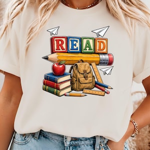 May include: A cream-colored t-shirt featuring a graphic print of colorful alphabet blocks spelling the word READ, a large yellow pencil, a red apple, a stack of three books, a brown backpack, two rulers, two pencils, and three paper airplanes.
