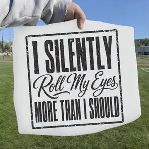 May include: A white square fabric item featuring a black distressed-style border and the black text "I SILENTLY Roll My Eyes MORE THAN I SHOULD" printed in a combination of bold block lettering and a cursive script font centered on the surface.