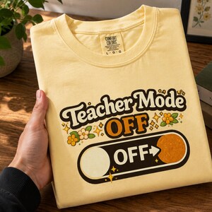 Puede incluir: Una camiseta amarilla clara con la frase "Teacher Mode OFF" en una fuente retro. El diseño incluye un gráfico de interruptor con la palabra "OFF" y detalles florales. La camiseta está doblada y sostenida por una mano.