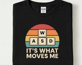 May include: Black t-shirt with a retro design featuring the letters "W", "A", "S", and "D" in a keyboard layout, set against a sunset-style background. The text "IT'S WHAT MOVES ME" is below the design.