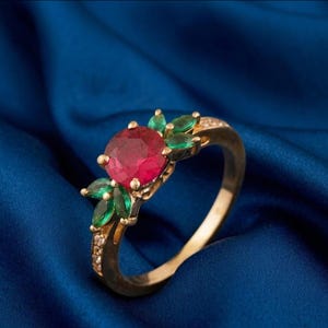 Puede incluir: Un anillo de oro con una gran gema roja redonda como punto focal, rodeada de gemas verdes más pequeñas y detalles delicados. La banda del anillo está adornada con pequeñas piedras transparentes.