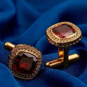 May include: A pair of gold cufflinks featuring square, deep red gemstones. Each cufflink has a border of small, clear stones. The cufflinks are presented on a blue, textured fabric.