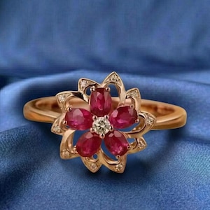 Puede incluir: Un anillo dorado con un diseño floral. La flor está compuesta por cinco piedras preciosas rojas oscuras de corte ovalado y una piedra preciosa clara redonda en el centro. Pequeñas piedras preciosas claras están engastadas alrededor de los pétalos. El anillo está sobre un fondo azul.