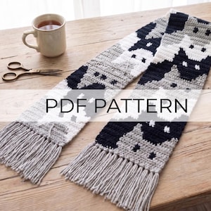 Peut inclure: Une longue écharpe au crochet présentant un motif répétitif de visages de chats en laine noire, blanche et grise. L'écharpe possède des franges gris clair aux deux extrémités. Le texte PDF PATTERN est superposé au centre en lettres capitales.