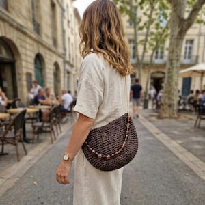 May include: A dark brown crocheted crescent-shaped shoulder bag with a textured woven pattern. The bag features a decorative strap adorned with translucent amber-coloured beads and a thin gold-tone chain, worn over a light beige linen-blend dress.