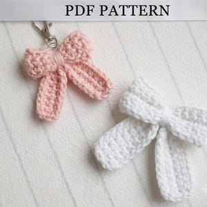 Pode incluir: Uma faixa com o texto PDF PATTERN acima de dois laços de crochê, um rosa claro e um branco. O laço rosa possui um fecho lagosta de metal prateado preso no centro superior, enquanto o laço branco não possui nenhum acessório.