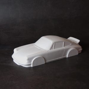 Può includere: Un'auto modello bianca, probabilmente un'auto sportiva classica, con uno spoiler posteriore. L'auto è un oggetto solido con una finitura liscia. L'immagine è ambientata su uno sfondo scuro.