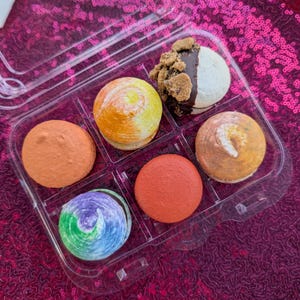 May include: A clear plastic container holding six colorful macarons. The macarons are various colors, including orange, yellow, brown, red, and a swirl of blue, green, and purple. One macaron is half-dipped in chocolate and topped with crumbs.