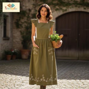 May include: An olive green apron with white floral embroidery on the pockets and hem. The apron has ruffled cap sleeves and a square neckline. A small basket of herbs and a loaf of bread are held in the hands.