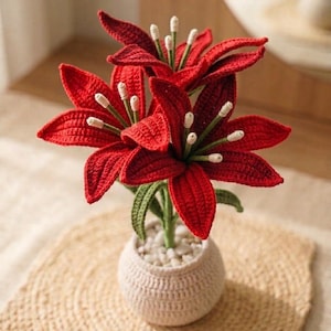 May include: A crochet lily plant featuring vibrant red petals with green stems and white-tipped stamens. The flowers are arranged in a small, round, cream-colored crocheted pot filled with white pebbles, creating a detailed textile floral display.