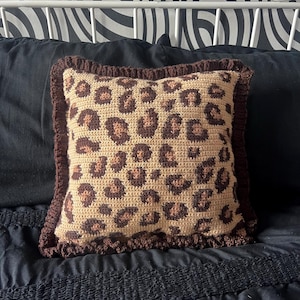 May include: A square crochet cushion featuring a leopard print design in tan and brown hues. It has a ruffled brown border and rests on a black bedspread. The background includes a white bed frame and a black and white patterned wall.