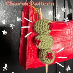May include: A crochet olive bag charm with three green olives and red accents, attached to a gold keyring. The charm is displayed against a red handbag. The text "Crochet Olive Bag Charm Pattern" is visible.