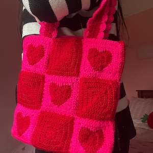 Peut inclure: Sac fourre-tout crocheté à la main, rose vif et rouge. Le sac présente un motif patchwork avec des cœurs rouges sur un fond rose. Les sangles et les bords sont également roses, créant un accessoire harmonieux et accrocheur.