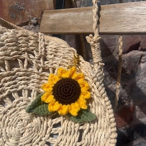 May include: A crocheted sunflower keychain with yellow petals and a brown center, attached to a woven straw bag. The bag has a circular design and a braided strap. The background includes a wooden chair and a brick wall.