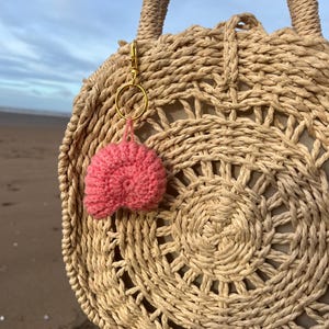 Könnte beinhalten: Eine runde, gewebte Strohtasche mit einem rosa Pompon-Schlüsselanhänger. Die Tasche hat ein kreisförmiges Design mit einem beigefarbenen Stofffutter. Der Pompon ist an einem goldfarbenen Ring und Verschluss befestigt. Der Hintergrund zeigt einen Strand und Himmel.