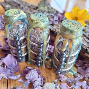 Può includere: Tre piccole bottiglie di vetro con tappi di sughero sigillati da cera verde e blu che cola. Ogni bottiglia è avvolta da uno spago e presenta una fila verticale di cinque perline grigio scuro. Contengono materiale vegetale essiccato.
