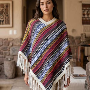 May include: A V-neck poncho featuring a multi-colored knit pattern with horizontal stripes in shades of red, yellow, blue, and black. The garment has a cream-colored trim along the neckline and hem, finished with long, cream-colored fringe tassels.