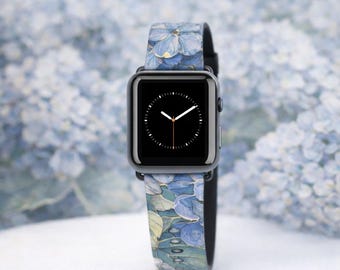May include: A black smartwatch with a floral patterned band featuring blue and green hydrangea blooms. The watch face displays a black screen with white hands. The background is a soft, blurred image of blue flowers.