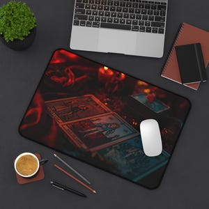May include: A rectangular mousepad featuring tarot cards with a mystical theme. The mousepad has a black border and a dark, reddish-brown background. A white computer mouse sits on the pad, near a laptop and other office supplies.