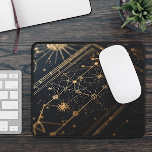 May include: Black rectangular mousepad with a gold celestial design featuring stars, constellations, and sunbursts. A white wireless mouse rests on the pad. The mousepad is on a dark wooden surface, with a keyboard and notebook visible.