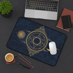 Puede incluir: Alfombrilla de escritorio rectangular con fondo azul oscuro y diseño oculto dorado. El diseño incluye un sol, una estrella y varios símbolos. Un ratón de ordenador blanco descansa sobre la alfombrilla.