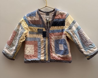 May include: A patchwork jacket in a variety of colors, including blue, yellow, and red, hangs on a gold hanger. The jacket features a quilted design with a V-neck and long sleeves. The jacket has a gray trim and the brand name "merenda" is visible.