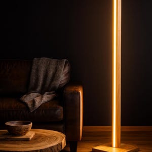 Può includere: Una moderna lampada da terra in legno con una striscia di luce calda al centro. La lampada è realizzata in legno chiaro e ha una base quadrata. La lampada è accanto a un divano in pelle marrone e un tavolino in legno.