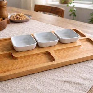 Puede incluir: Una bandeja de madera con tres cuencos cuadrados blancos y dos secciones empotradas. La bandeja es de madera clara y se encuentra sobre un mantel de color neutro. Otros cuencos de madera y saleros y pimenteros están en el fondo.
