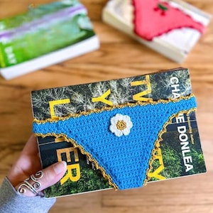 Peut inclure: Une housse de livre en crochet bleu avec une applique de fleur blanche au centre jaune et une bordure festonnée dorée. La housse entoure un livre intitulé The Girl Who Was Taken de Charlie Donlea, avec une housse verte et une rose visibles.