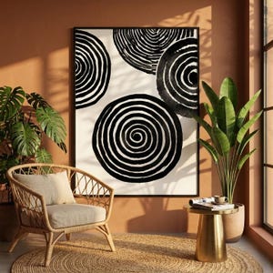 Peut inclure: Une impression d'art encadrée avec des cercles concentriques noirs et blancs. L'œuvre est exposée dans une pièce avec un fauteuil en rotin, un tapis rond en jute et une table d'appoint dorée. Une plante en pot est près de la fenêtre.