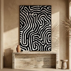 Peut inclure: Impression d'art abstrait en noir et blanc dans un cadre noir. L'œuvre présente un motif audacieux de lignes épaisses et courbes. L'impression est au-dessus d'une console en pierre avec un petit vase et des livres.