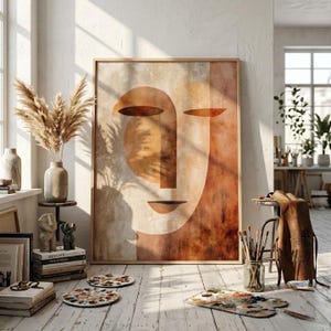 Puede incluir: Gran impresión artística abstracta enmarcada con un diseño de rostro minimalista en tonos marrones y beige. La obra de arte se exhibe en una habitación luminosa con una estética rústica, junto con objetos decorativos y materiales de arte.